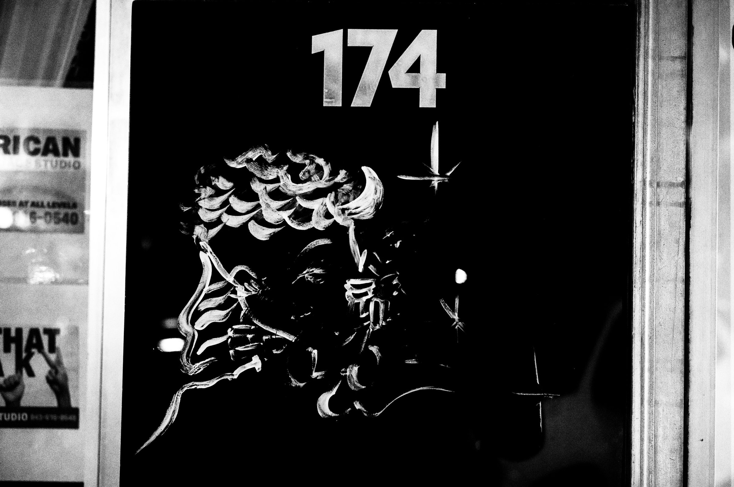 High-contrast black-and-white photograph of a dark glass door marked “174,” with a loosely painted white line drawing of a curly-haired figure in profile and small star-like sparkles.