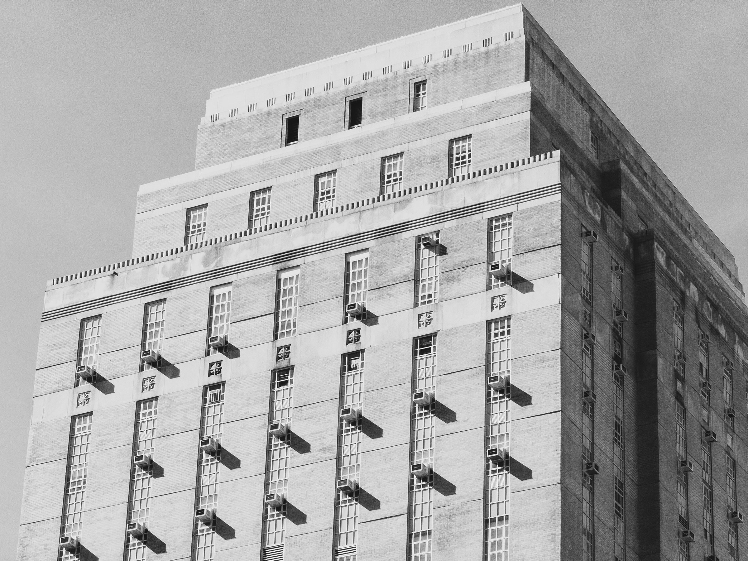 Black-and-white photograph looking up at the stepped upper floors of a brick Art Deco high-rise against a plain gray sky. Tall, narrow multi-pane windows run in vertical columns, and window air conditioners project from many of them, each casting a long diagonal shadow onto the sunlit brick. Small carved ornaments sit between the window columns, and the setbacks are topped with tooth-like cornice detailing. The right side of the building is in deep shade.