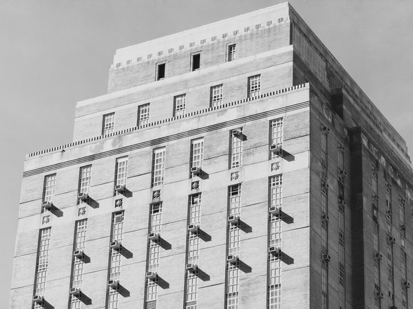 Black-and-white photograph looking up at the stepped upper floors of a brick Art Deco high-rise against a plain gray sky. Tall, narrow multi-pane windows run in vertical columns, and window air conditioners project from many of them, each casting a long diagonal shadow onto the sunlit brick. Small carved ornaments sit between the window columns, and the setbacks are topped with tooth-like cornice detailing. The right side of the building is in deep shade.