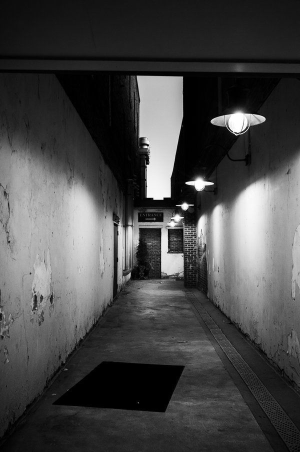 Black-and-white photograph of a narrow, dimly lit alley with peeling plaster walls, a row of glowing barn-style lamps on the right, and an “ENTRANCE” sign with an arrow at the far end.
