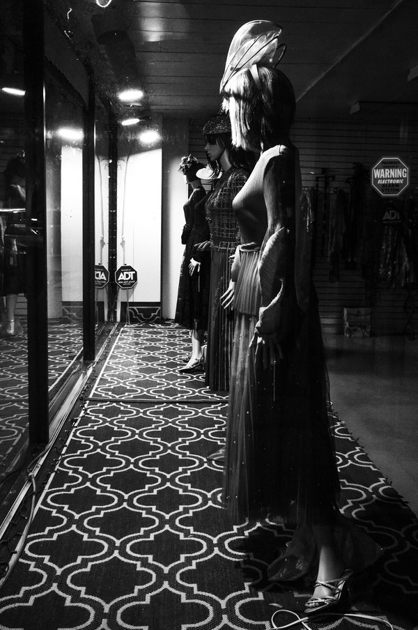 Black-and-white photograph of three mannequins in elegant gowns and hats, lined up in a shop window on a geometric patterned rug, with an ADT security sign in front of  them and a WARNING sign behind them.