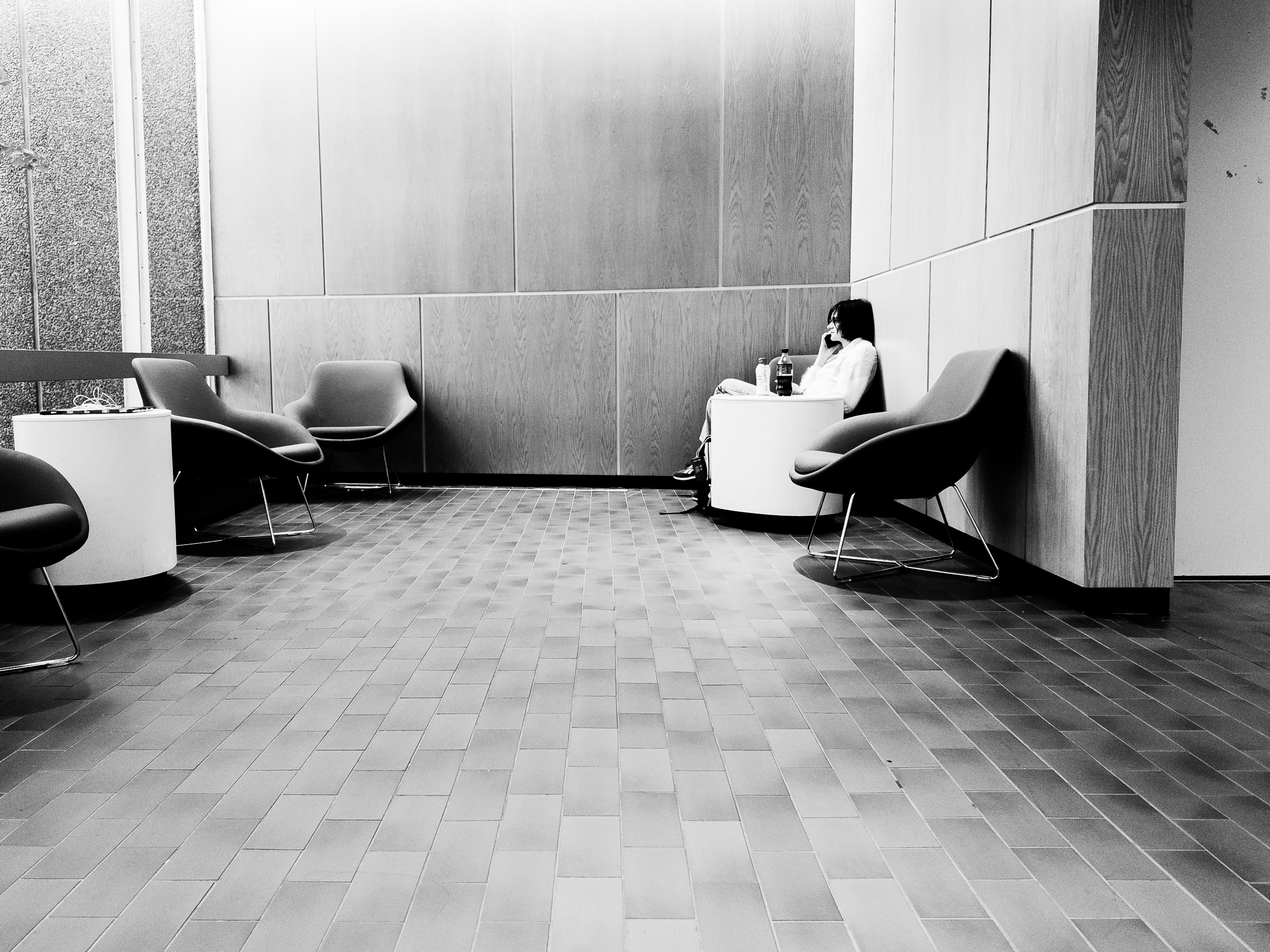 Black-and-white photograph of a spacious lounge with wood-paneled walls and a floor of rectangular tiles. In the upper right, a small figure with short dark hair sits in a corner holding a phone to their ear, with two bottles on a round white table in front of them. Modern bucket chairs on thin metal legs sit around the room, including two beside another round table on the left. Most of the frame is the empty tile floor and large panels of the walls, emphasizing the isolating effect of talking on a phone in a public space.