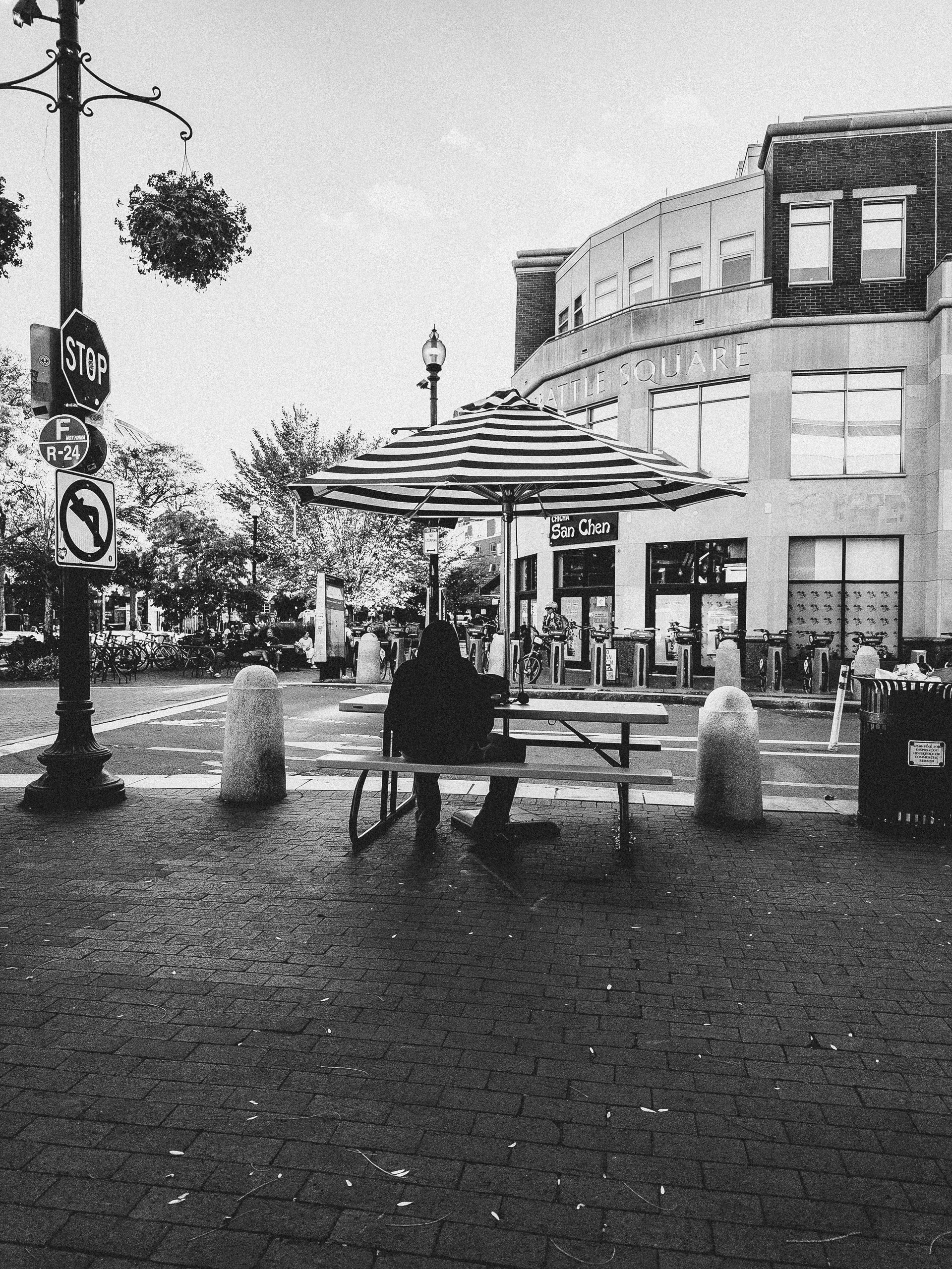 Black-and-white street photograph of a person in a dark hooded top seen from behind, sitting alone at a picnic table beneath a striped umbrella on a brick plaza. In the foreground, a lamppost carries a stop sign and a no-left-turn sign, with a hanging flower basket above. Stone bollards line the edge of the plaza, and a row of bike-share docks stands in front of a curved building with “Battle Square” lettered on its facade. Trees and a few seated people fill the background, and a trash bin sits at the right edge.
