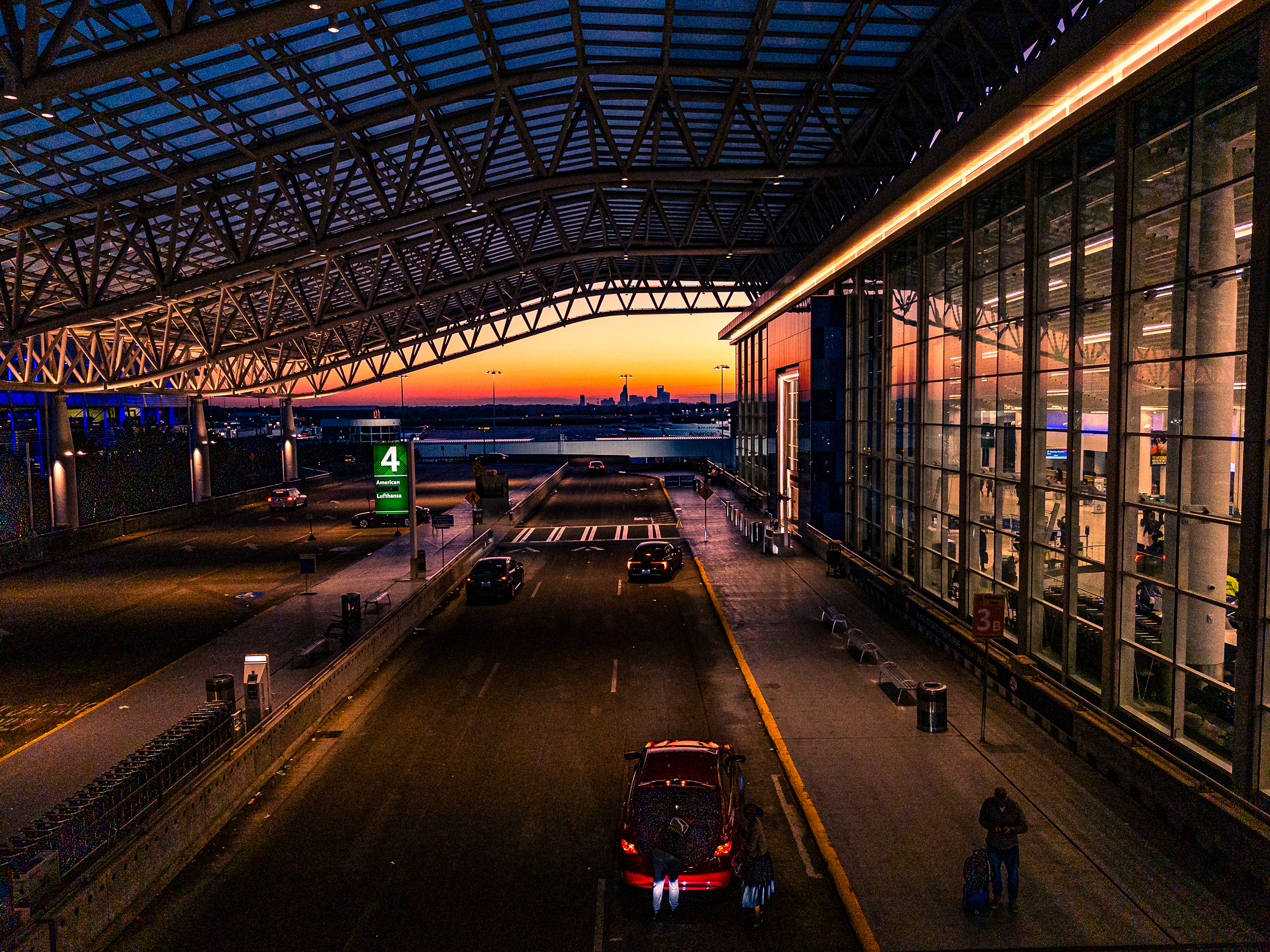 Sunrise view from an elevated walkway looking along the road adjacent the terminal at Charlotte Douglas International Airport, framed by a curved steel-truss canopy, with an orange-to-blue sky and the Charlotte skyline in the distance.