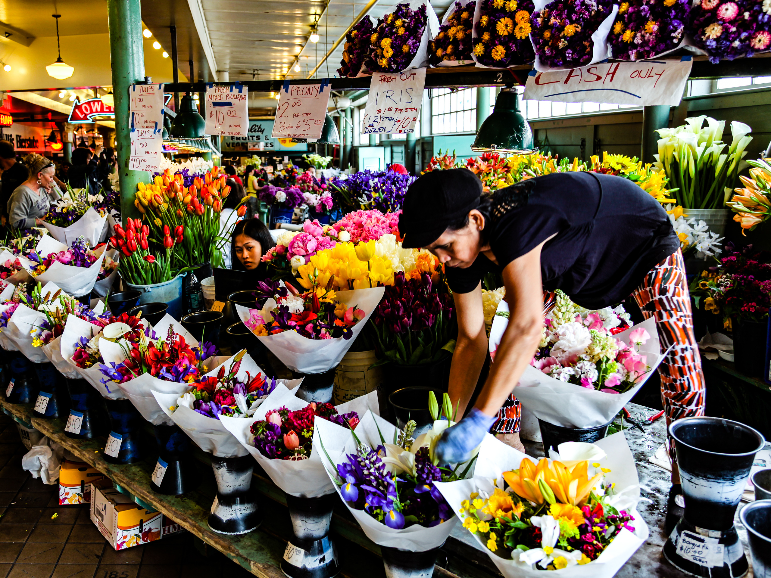 A woman in a black cap and blue gloves leans over a crowded flower stall, arranging white-wrapped bouquets of tulips, irises, peonies and lilies in saturated reds, purples, pinks and yellows, with handwritten price signs and a “CASH ONLY” sign hanging overhead. A female assistant looks on in the background.