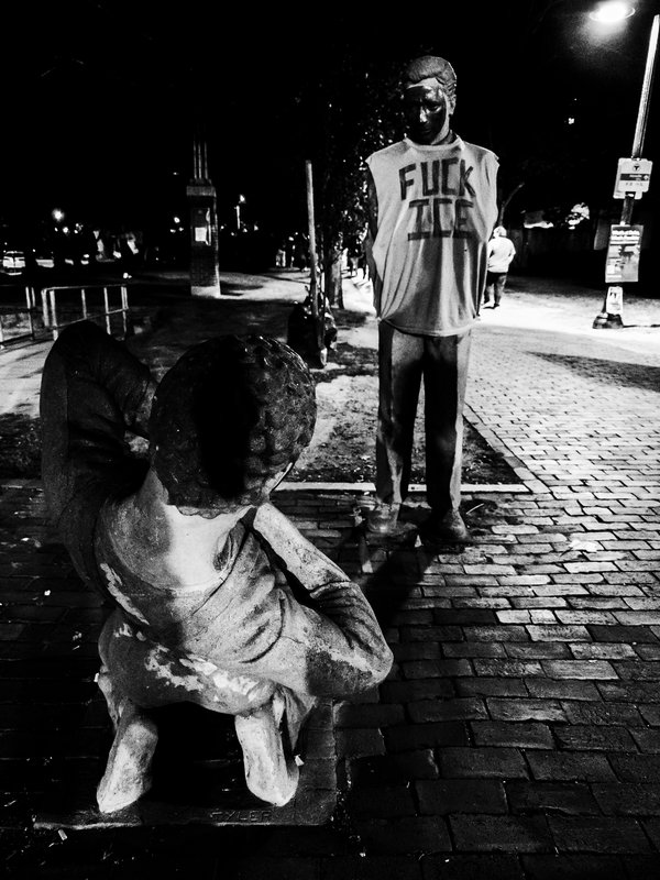 Black-and-white night photo of two bronze sculptures on a brick pedestrian path: a figure crouched in the foreground, seen from behind, faces a standing figure with concealed arms and accesorized with a sleeveless shirt hand-lettered “FUCK ICE.”