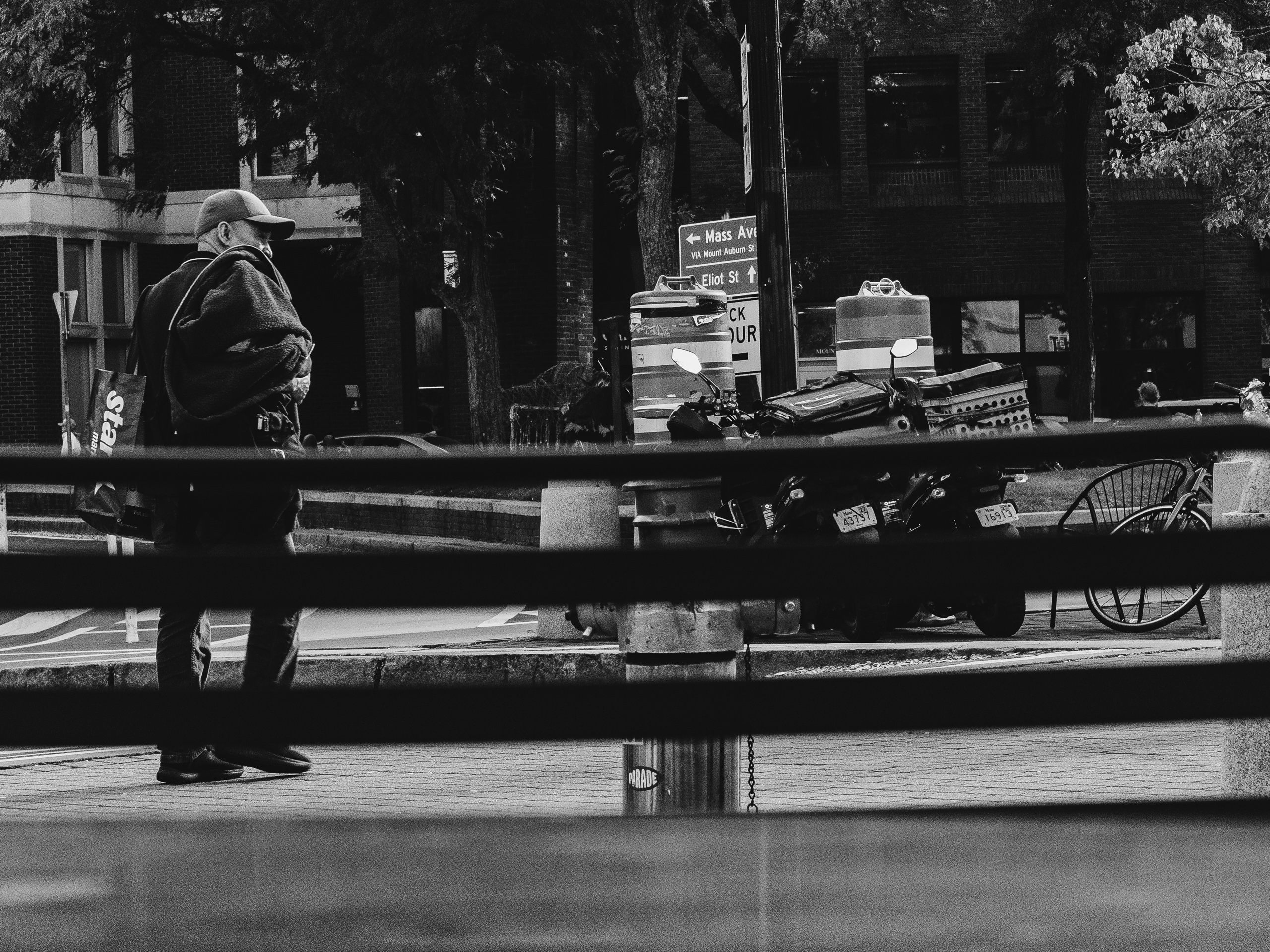 Black-and-white street photograph taken through dark horizontal railings of a bench back. On the left, a man in a baseball cap looks down and to the side while carrying a bulky fleece jacket over his shoulder and a shopping bag with partial white lettering. Behind him, a street sign points toward Mass Ave and Eliot St. Two construction barrels stand beside a two parked delivery scooters. A bicycle and a wire chair sit to the right, and dark brick buildings and leafy trees fill the background.