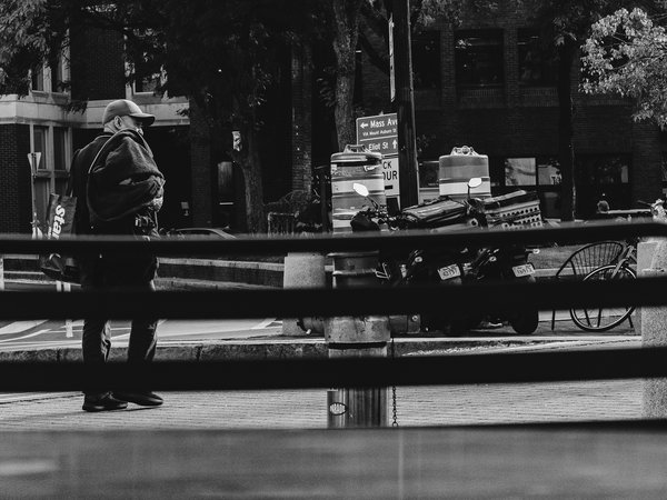 Black-and-white street photograph taken through dark horizontal railings of a bench back. On the left, a man in a baseball cap looks down and to the side while carrying a bulky fleece jacket over his shoulder and a shopping bag with partial white lettering. Behind him, a street sign points toward Mass Ave and Eliot St. Two construction barrels stand beside a two parked delivery scooters. A bicycle and a wire chair sit to the right, and dark brick buildings and leafy trees fill the background.