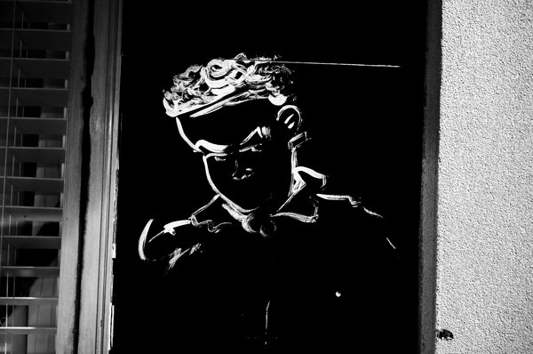 High-contrast black-and-white photograph of a white line drawing of a stern, curly-haired figure painted on a black glass door, set beside a textured stucco wall.
