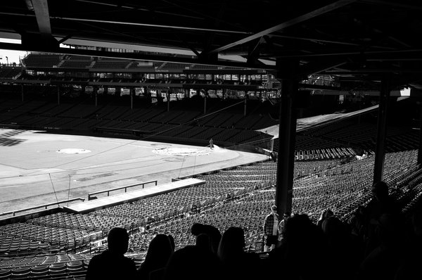 Black-and-white view from beneath a dark steel-framed grandstand roof, looking down on an empty ballpark. Sunlight rakes across thousands of vacant seats and the infield. A lone groundskeeper waters the grass near home plate. Silhouetted tourists fill the bottom foreground, and a man in a plaid shirt and sunglasses stands in the aisle beyond them.
