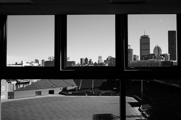 Black-and-white photograph taken from a dark interior room. Three window panes frame a clear sky and the Boston skyline, including the Prudential Tower. Below the window frame, an empty outfield and rows of unoccupied bleacher seats stretch out in muted gray tones.