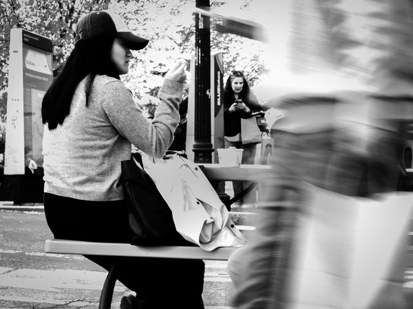 Black-and-white street photograph of a woman in a baseball cap and knit sweater sitting sideways on a picnic table bench, raising a hand toward her mouth as she eats. A jacket and dark bag rest on the bench beside her, and a takeout cup sits on the table. Behind her, a woman with wavy hair looks down at a phone near a Bluebikes station and a lamppost. On the right, a blurred figure moves through the foreground while carrying a shopping bag, and bright, leafy trees fill the background.