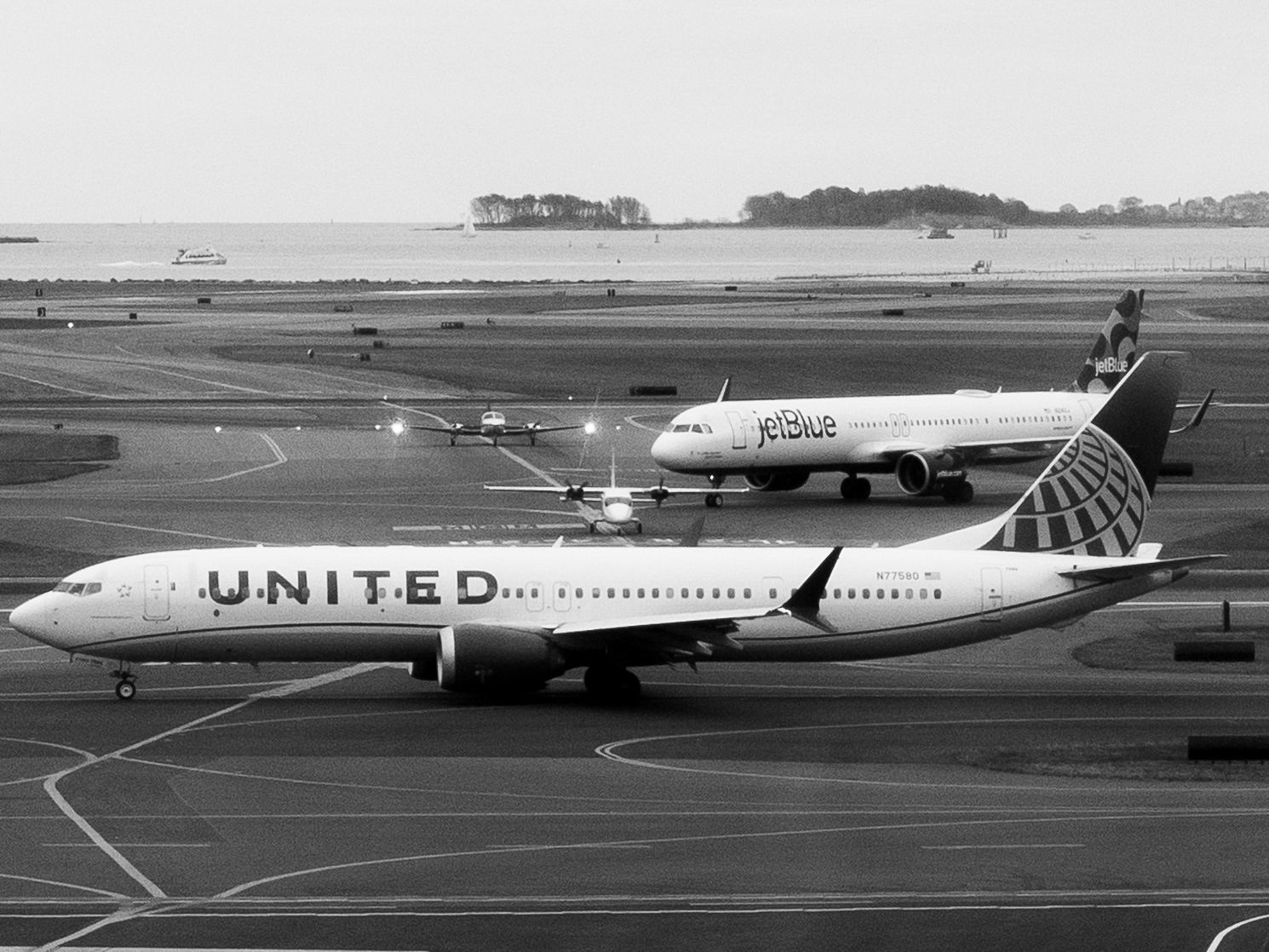 Black-and-white photo of a United Airlines Boeing 737 MAX taxiing left to right across an airport apron, with a JetBlue Airbus A321 and two smaller propeller planes on the taxiways behind it, and a harbor with wooded islands in the distance.