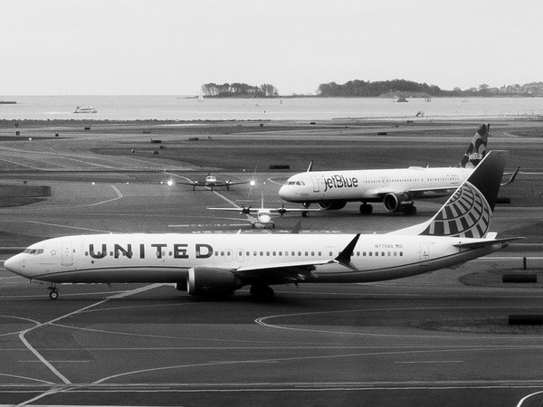 Black-and-white photo of a United Airlines Boeing 737 MAX taxiing left to right across an airport apron, with a JetBlue Airbus A321 and two smaller propeller planes on the taxiways behind it, and a harbor with wooded islands in the distance.