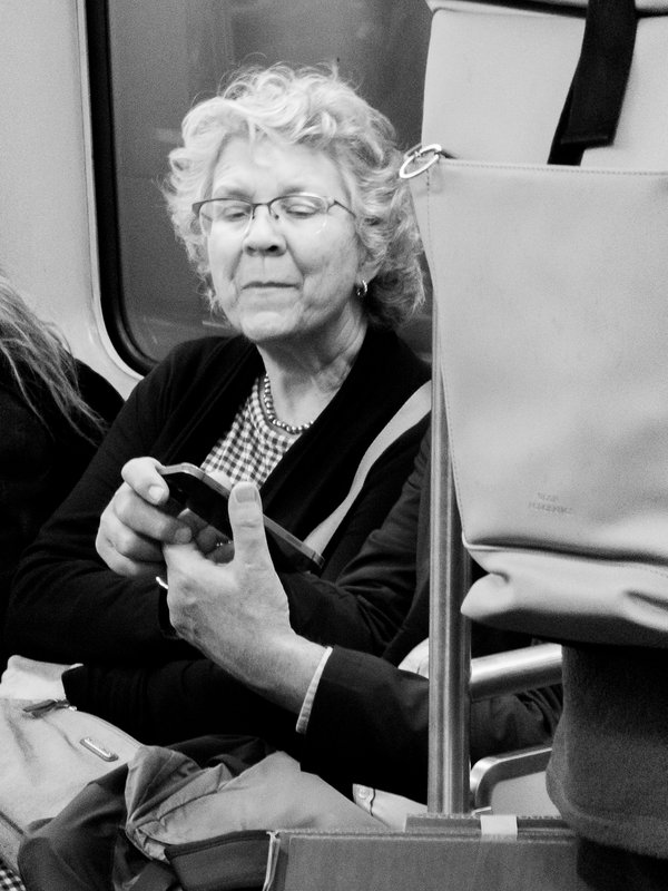 Black-and-white candid portrait of an elderly woman seated on a subway car, looking down at a smartphone held in hands of a passenger sitting next to her. She has short, curly light hair, wire-rimmed glasses, a beaded necklace, a gingham top and a dark cardigan. Her expression is a faint, amused smile. A metal pole runs vertically through the right of the frame beside a hanging tote bag, and a bag rests in her lap. The face of the person holding the phone is hidden by a backpack of another passenger.