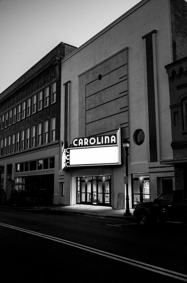 Black-and-white photograph of the Carolina Theatre at dusk, its Art Deco façade and glowing blank marquee standing out against a dark, empty street.
