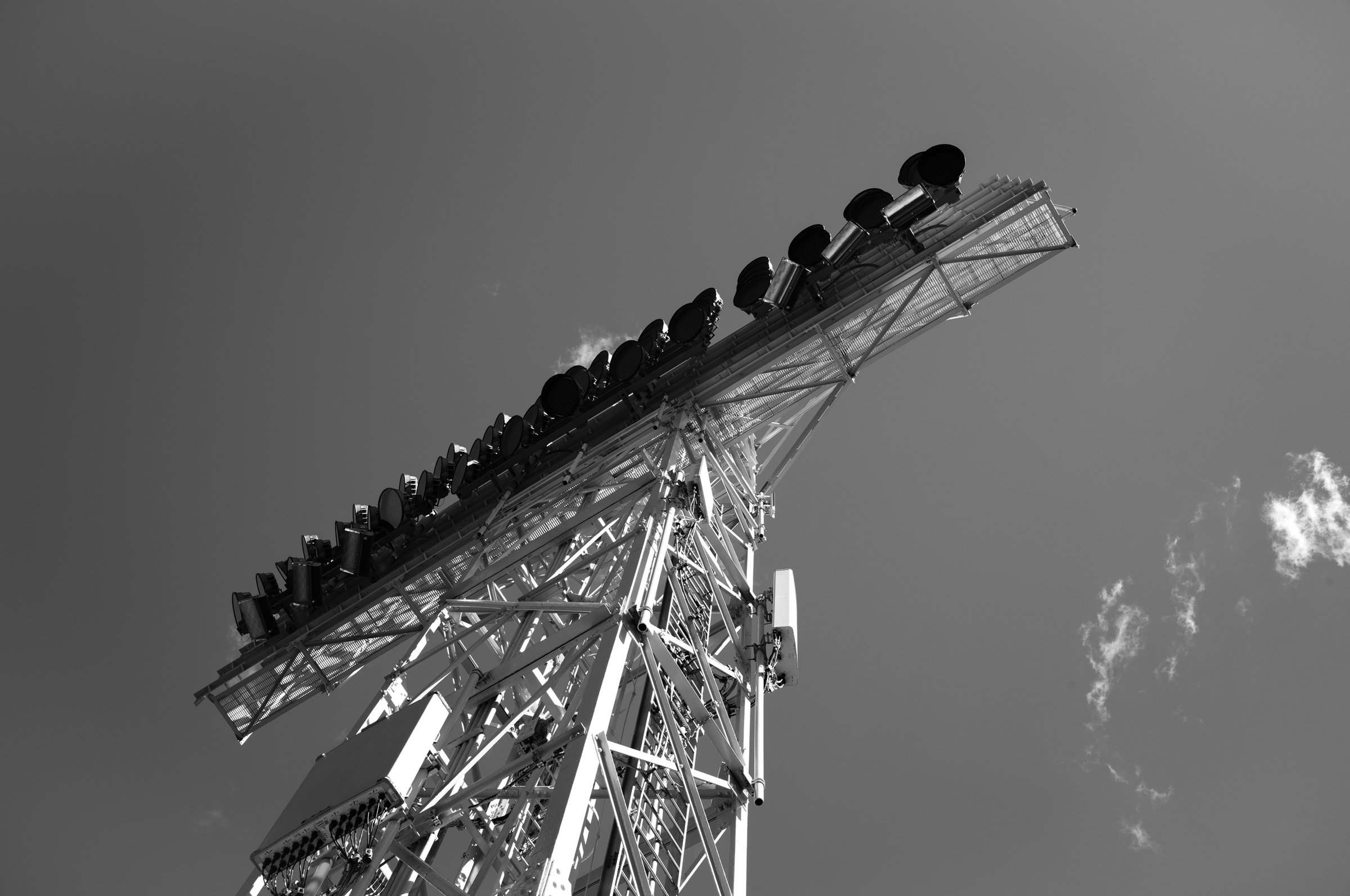 Black-and-white upward-angled shot of a white steel lattice tower topped with a catwalk and a row of dark floodlights. The tower slants diagonally across the frame against a gray sky with a few wispy clouds at right.